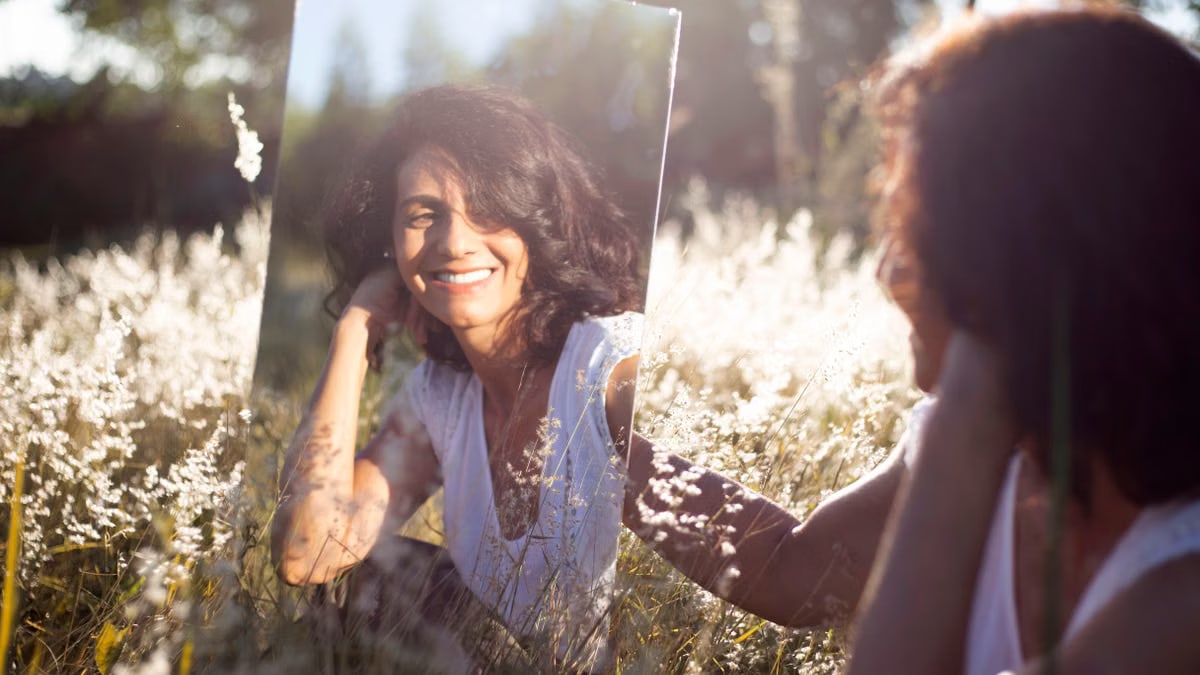 A woman smiles at her reflection in a mirror placed in a sunny field.
